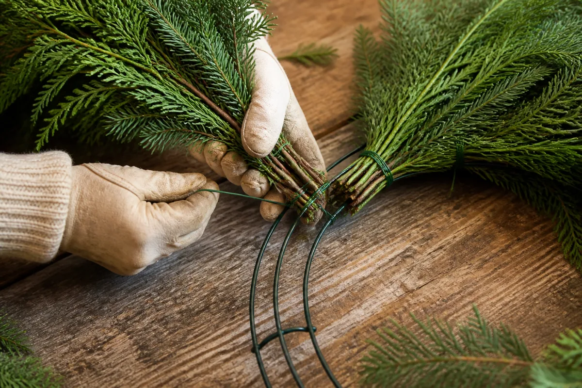 Gloved hands securing overlapping evergreen bundles to a wire wreath frame with green paddle wire